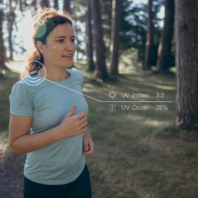 Woman wearing sun-a-wear UV tracker on her t-shirst collar while running through park. Information shows UV-Index of 3.2 and UV-Dose of 78 percent